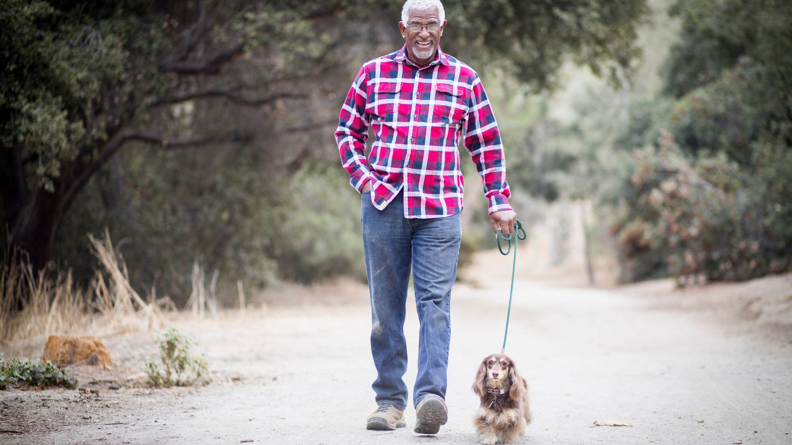 Older man in plaid shirt and jeans walking a dachshund down a country road